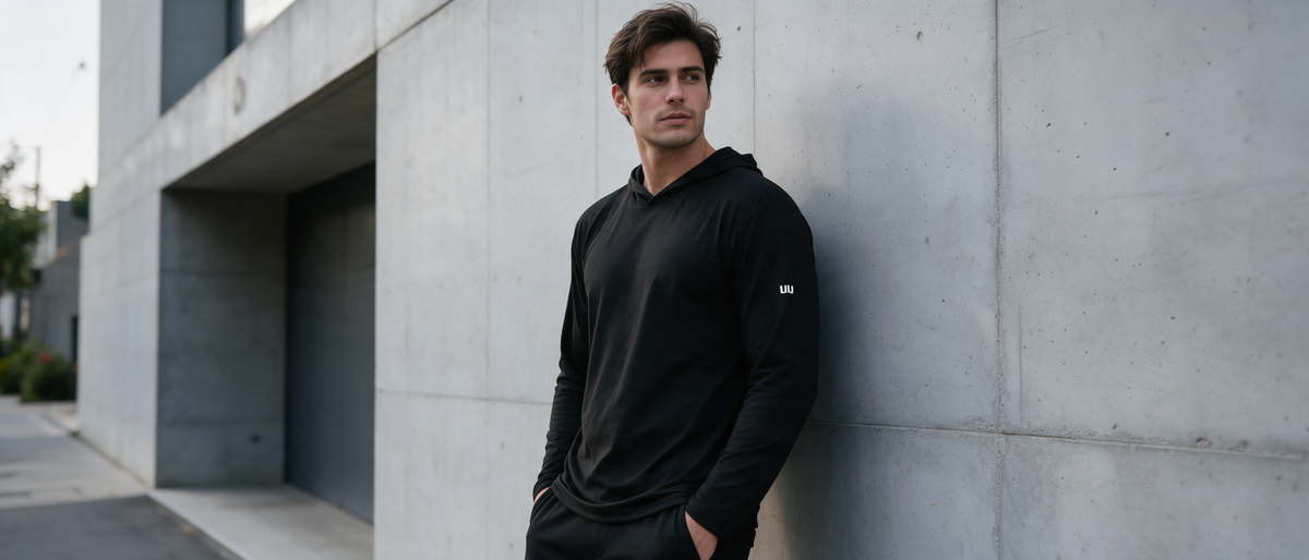 Hombre vistiendo sudadera negra TUUM Blizzard con capucha y logotipo blanco lateral en una sesión de moda urbana junto a una pared de concreto.