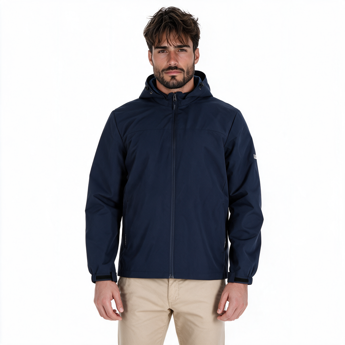 Hombre modelando chamarra TUUM Peak azul marino con capucha, cierre frontal y logotipo blanco en manga, combinada con pantalón beige sobre fondo blanco.
