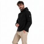 Modelo masculino luciendo chamarra TUUM Peak negra con capucha y cierre frontal, en pose editorial de tres cuartos con pantalón beige sobre fondo blanco.