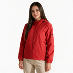 Mujer modelando chamarra TUUM Peak roja con capucha ajustable, cierre frontal y logotipo negro en manga, combinada con pantalón beige sobre fondo blanco